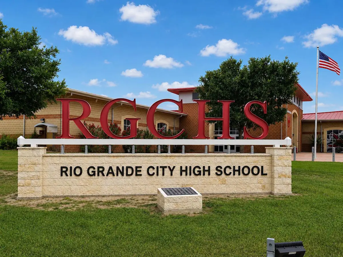 Front campus sign for Rio Grande City High School, with large red “RGCHS” letters above the school name, a tan school building, trees, lawn, and an American flag under a blue sky.