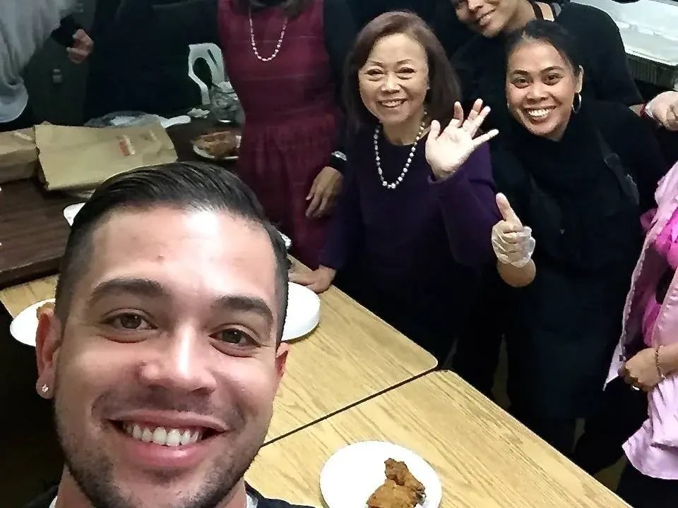 A group of students and adults pose around a table indoors, with lockers behind them; a person in the foreground holds the camera for a selfie.