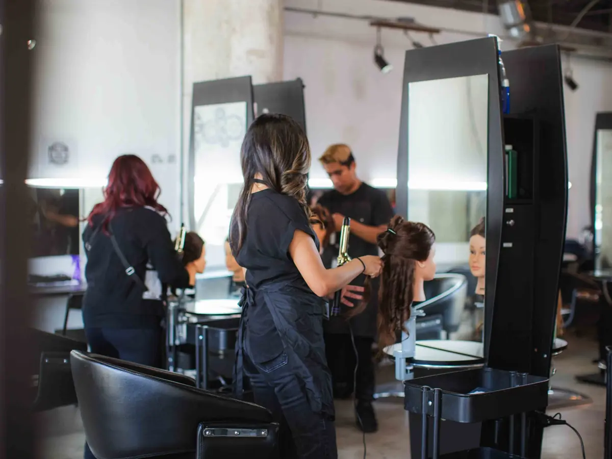 Students and instructors work at styling stations in a bright cosmetology classroom, with mirrors, salon chairs, and hair-washing equipment visible.