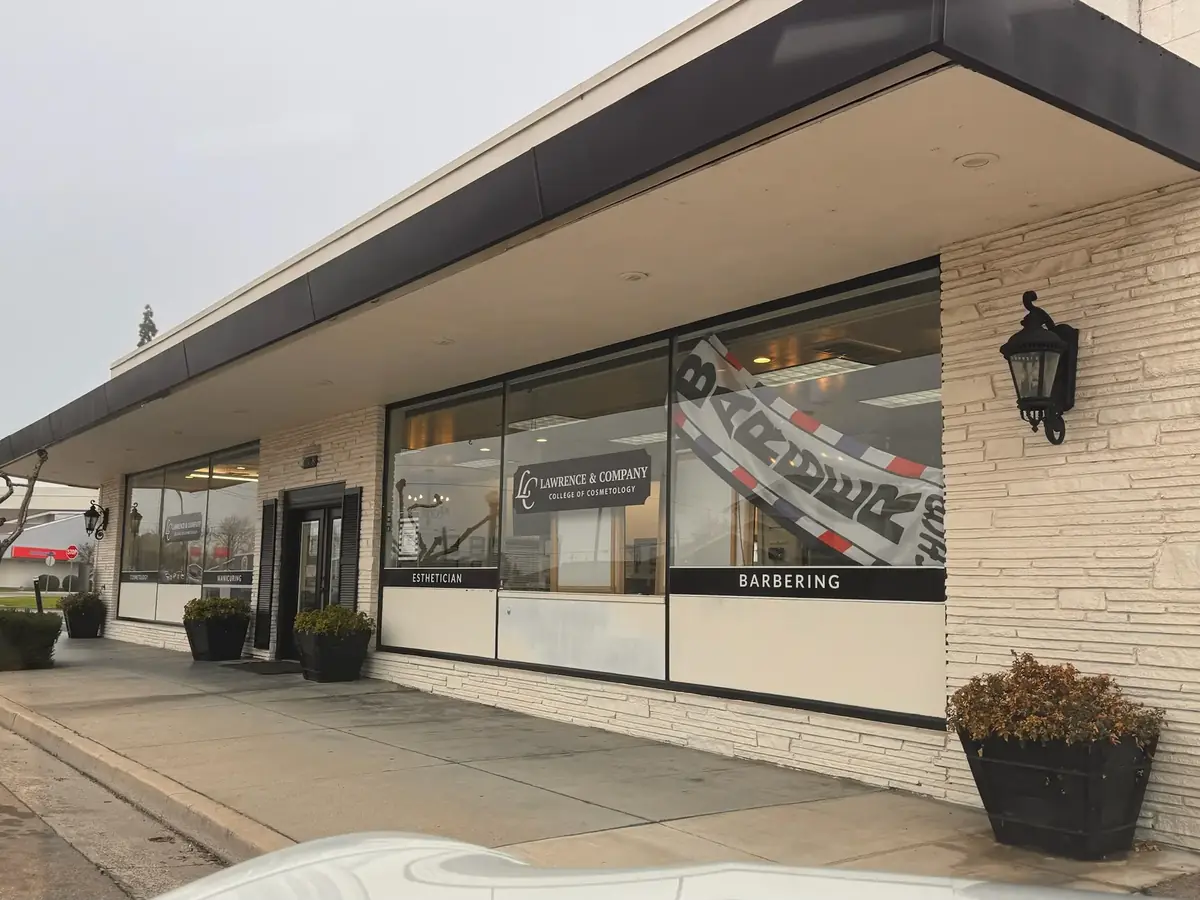 Exterior of Lawrence & Company College of Cosmetology in Selma, California, showing the white brick storefront, large windows with cosmetology and barbering signage, sidewalk, and planters.