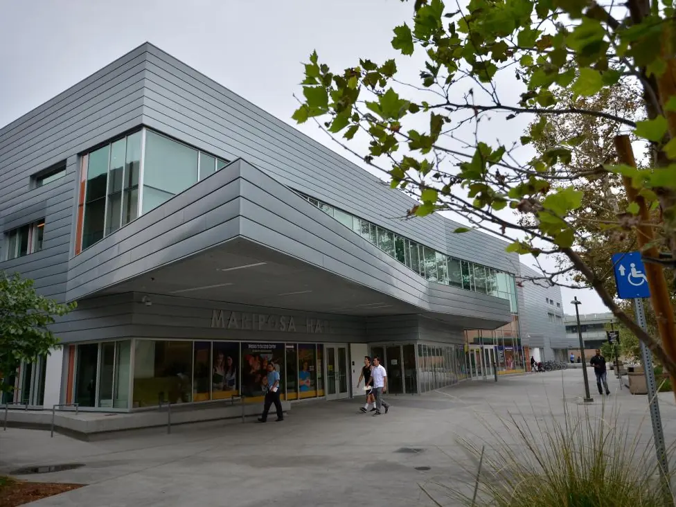 Exterior of a modern Los Angeles Trade Technical College campus building with a covered entrance, glass windows, trees, accessible parking signage, and people walking through the plaza.