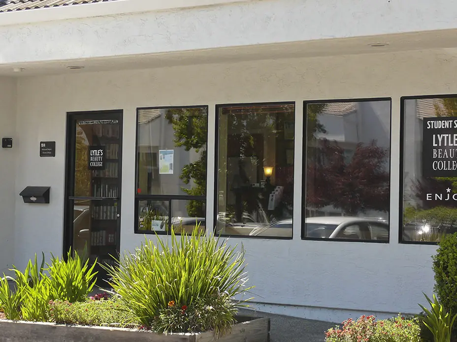 Exterior of Lytle’s Beauty College student salon in a white building, with dark windows displaying the school name and landscaped plants along the entrance.