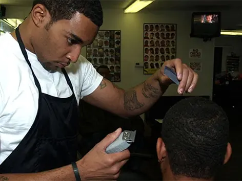 A barber in a white shirt and black apron trims a seated client's hair with electric clippers inside a barber-training classroom, with haircut posters and mirrors in the background.