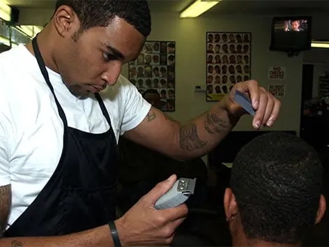A barber in a white shirt and black apron uses electric clippers on a seated client's hair inside a barber training salon.