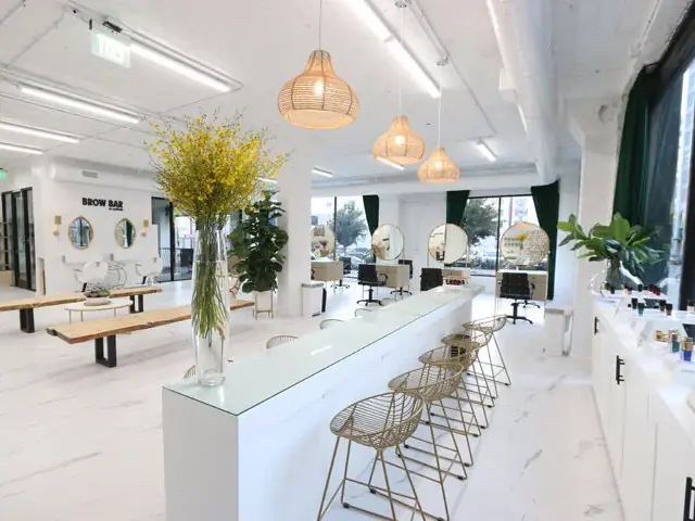 Bright white beauty academy salon interior with a long central counter, wire bar stools, round salon mirrors, styling stations, pendant lights, plants, and large windows.
