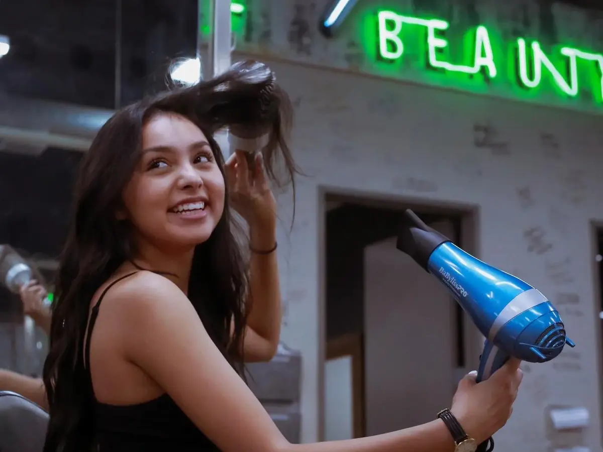 Student holding a blue hair dryer while styling a seated client's hair inside the Newberry School of Beauty salon, with illuminated Newberry Beauty signage behind them.