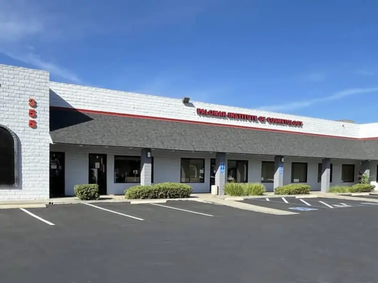 Exterior of the Palomar Institute of Cosmetology building with its name displayed above the entrance, a covered walkway, parking lot, and blue sky.