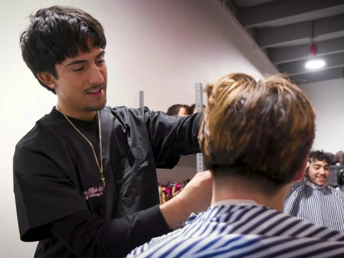 A student in a black barbering cape trims the hair of a seated client wearing a striped salon cape, with other students and salon stations visible in the background.