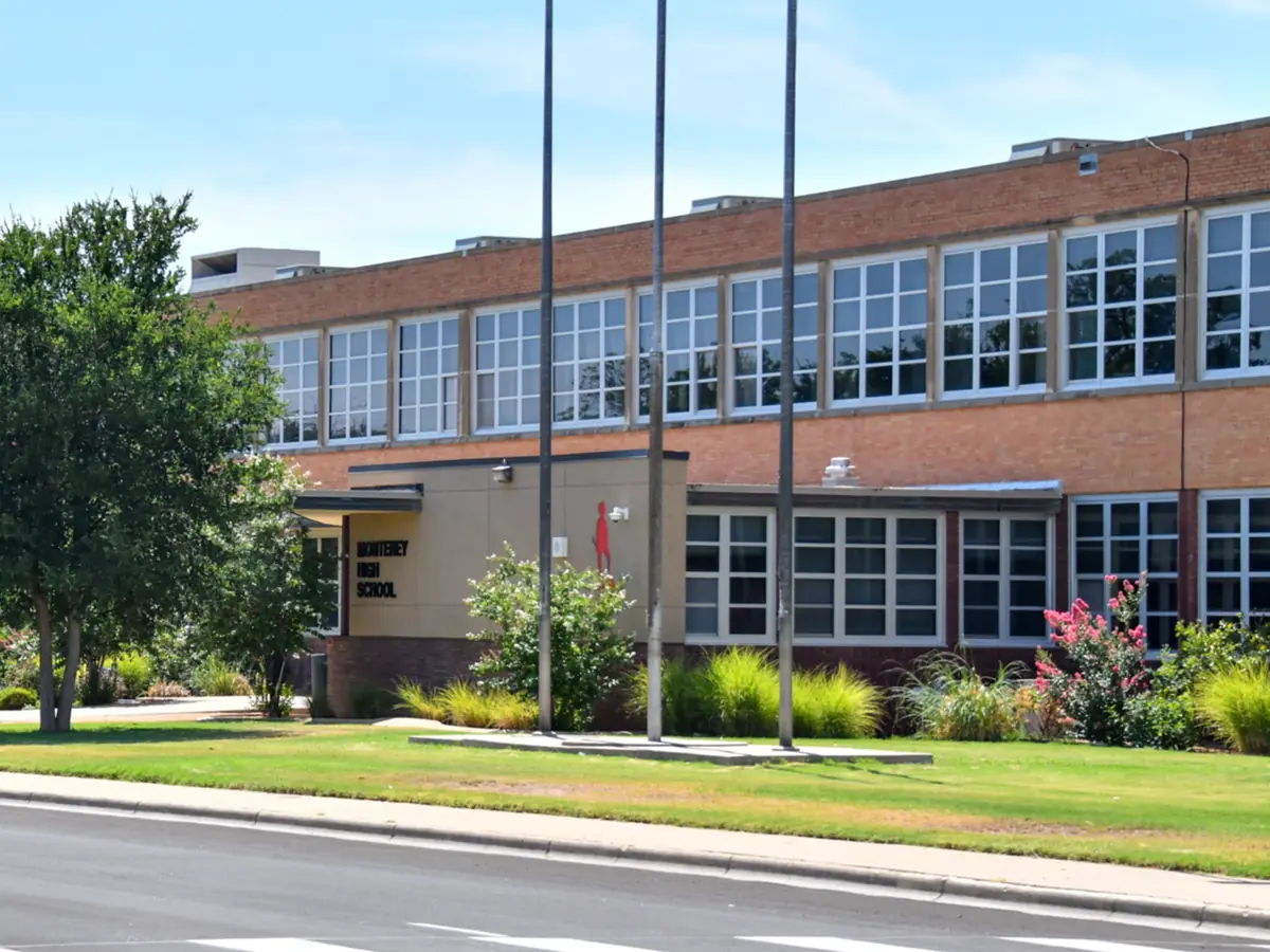 Two-story red-brick Monterey High School building with large rows of windows, flagpoles, trees, landscaped grounds, and a street in front.