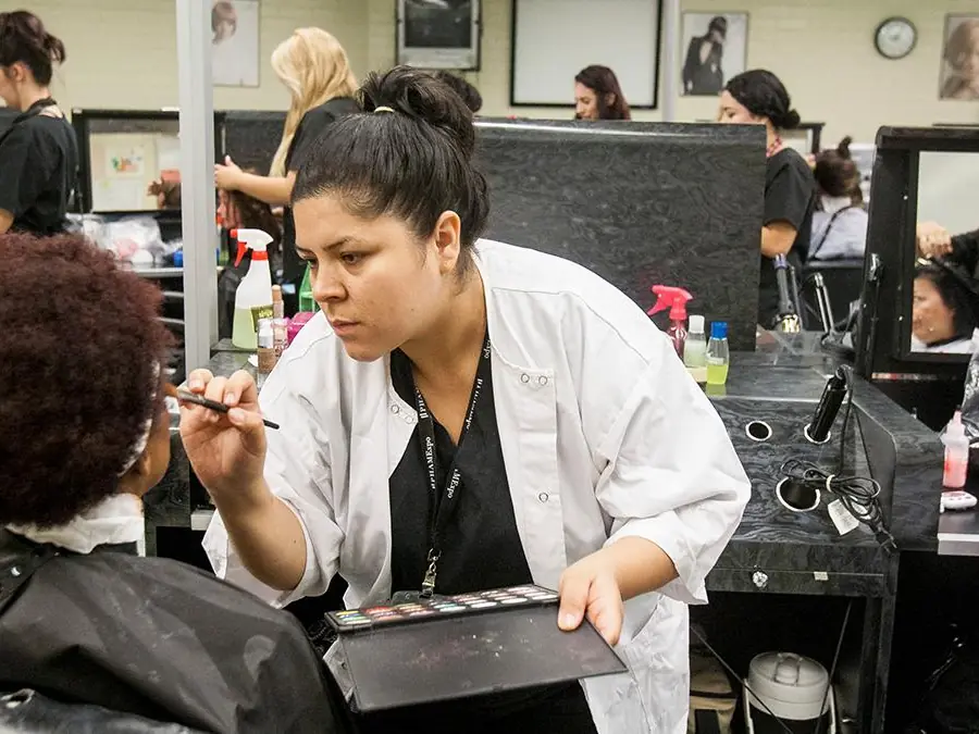 A cosmetology student in a white coat applies makeup to a seated client in an RCC salon, with mirrors, styling stations, and other salon workers visible behind them.