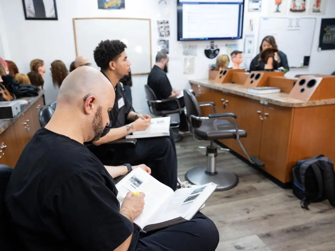 Students seated in a cosmetology classroom with salon chairs and workstations while an instructor presents at the front of the room.