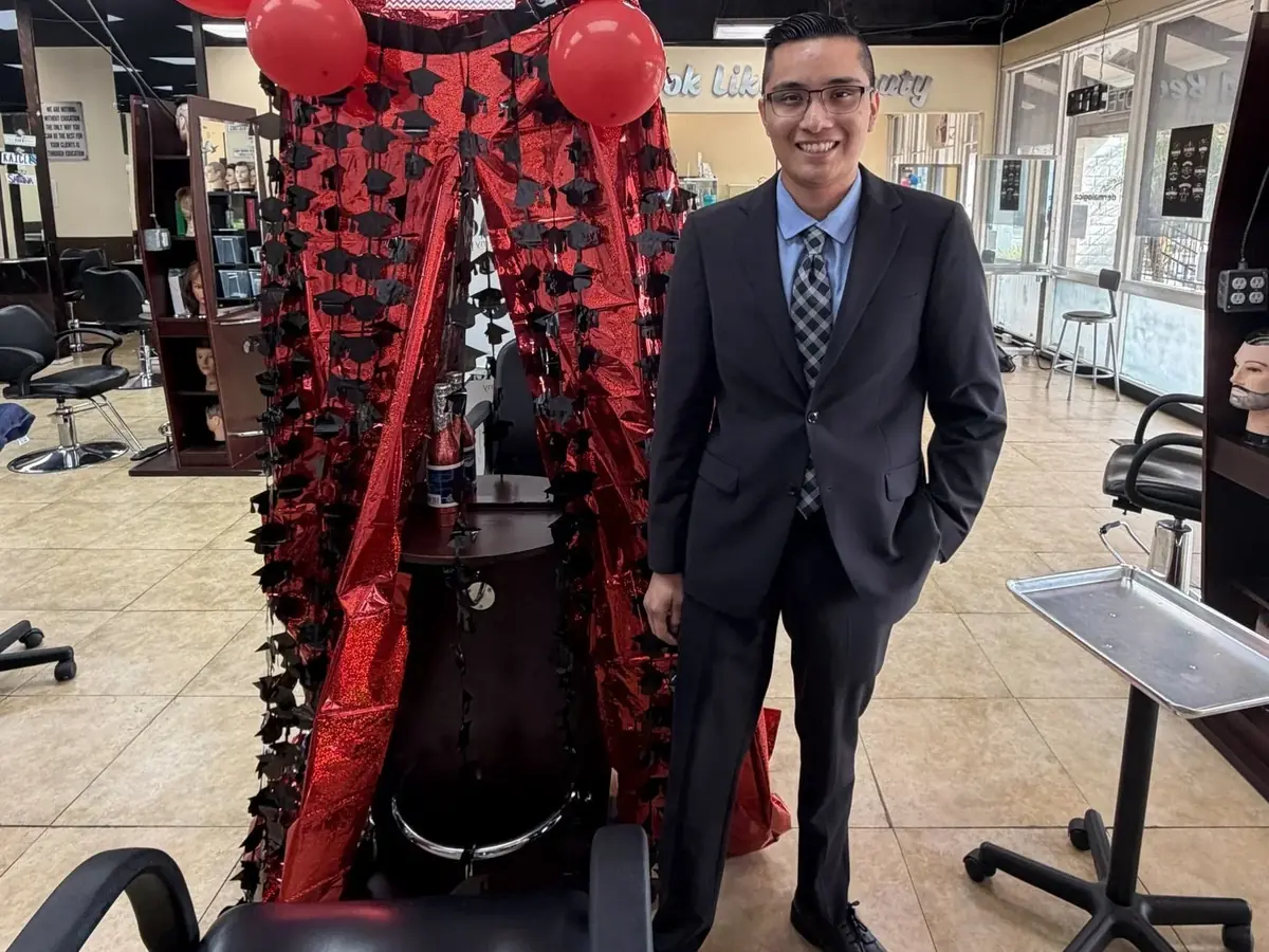 A graduate in a dark suit stands inside Saddleback Beauty Academy’s salon beside a decorated barber chair with red balloons and a red-and-black backdrop; salon stations and styling chairs are visible around him.