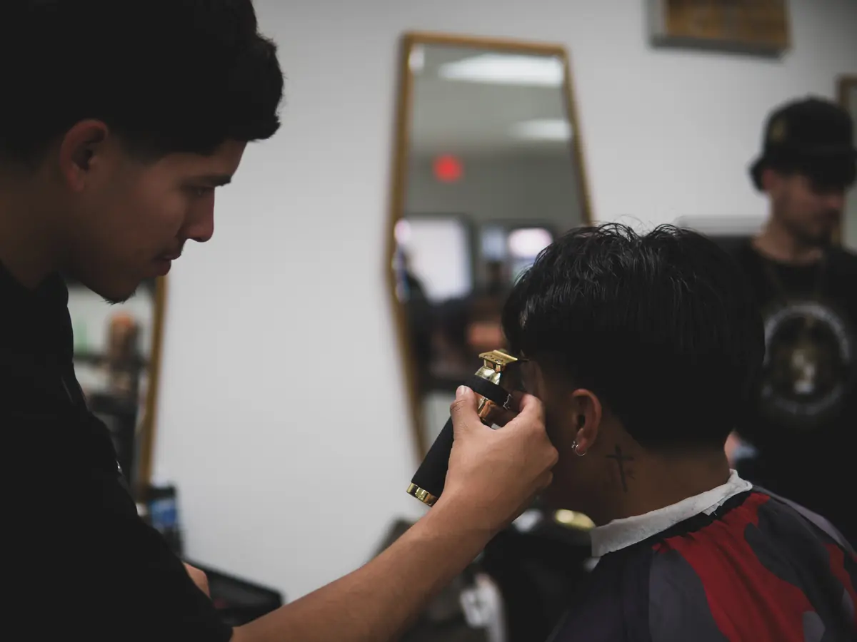 A barbering student uses electric clippers to cut a seated client’s hair in a salon classroom, with mirrors and another person visible in the background.