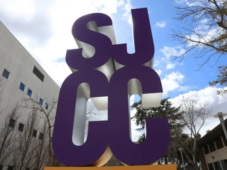 Large purple SJCC sculpture standing between campus buildings under a partly cloudy sky.