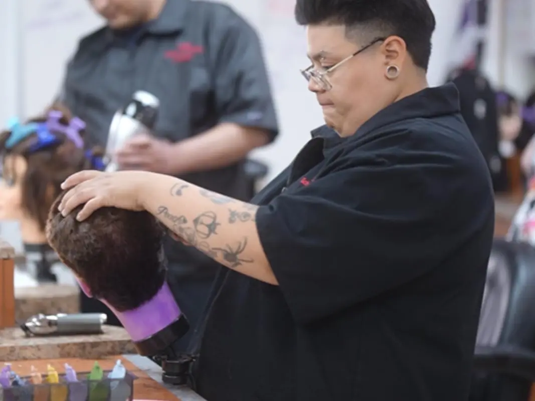 Student in a black uniform styles a mannequin head at a salon workstation while other students practice in the background.