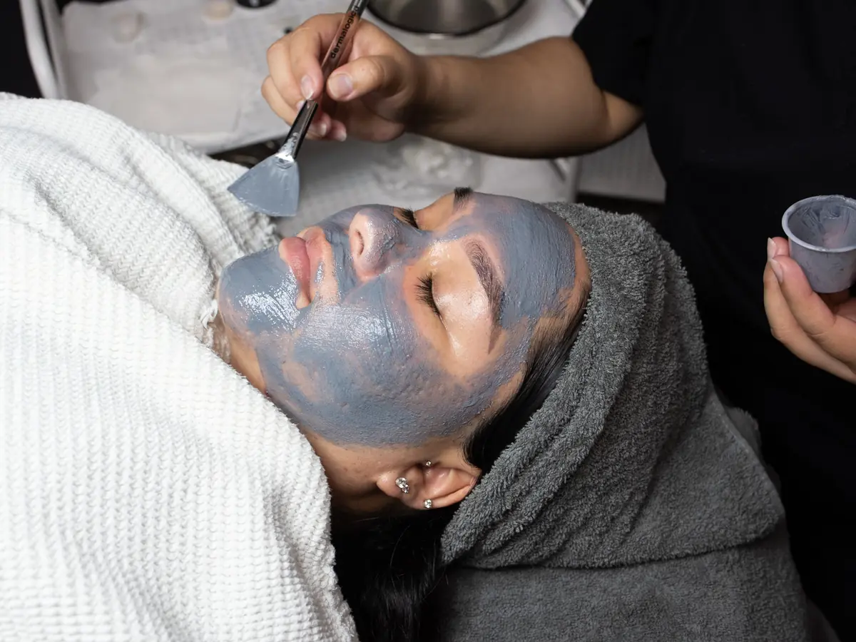 A person receives a gray facial mask treatment while lying under a white towel, as another person applies the mask with a brush.