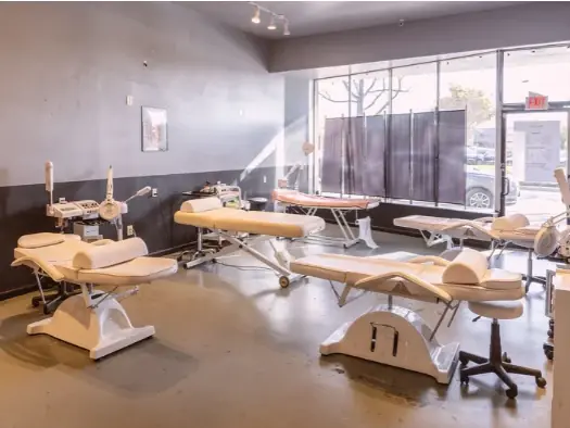Esthetics classroom with several cream-colored treatment tables and equipment arranged in a bright room beside large front windows.