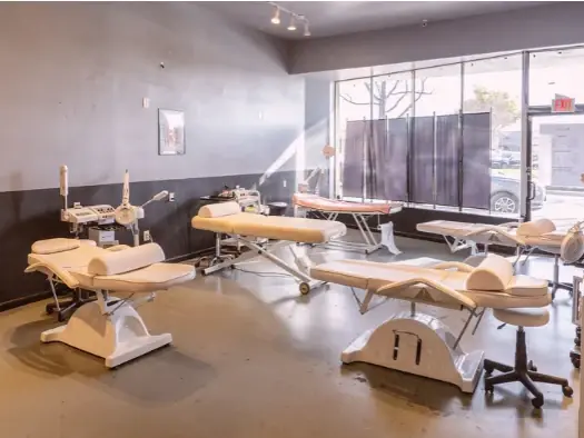 Bright esthetics classroom with several cream-colored treatment tables and equipment arranged across a polished floor, with large front windows.