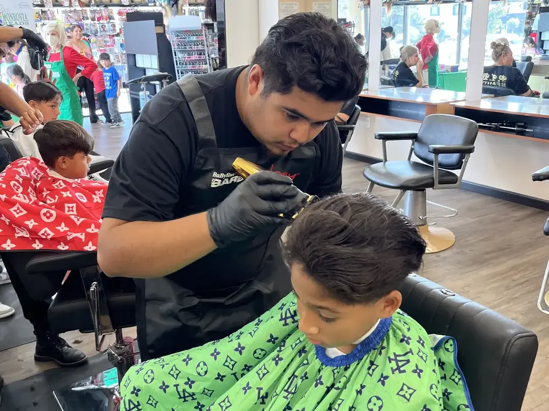 A student barber wearing black gloves cuts a seated client's hair in a bright salon, with barber chairs, mirrors, hair products, and other haircutting stations visible around them.